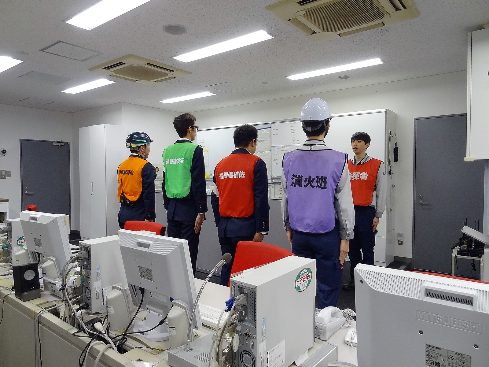 防災センターで、パソコンが並んだデスクを背に、「消化班」などの文字が書かれた紫、赤、緑、オレンジのカラーベストを着た男性たちが、「指揮者」のベストを着た男性から指示を受けている、訓練の様子の写真