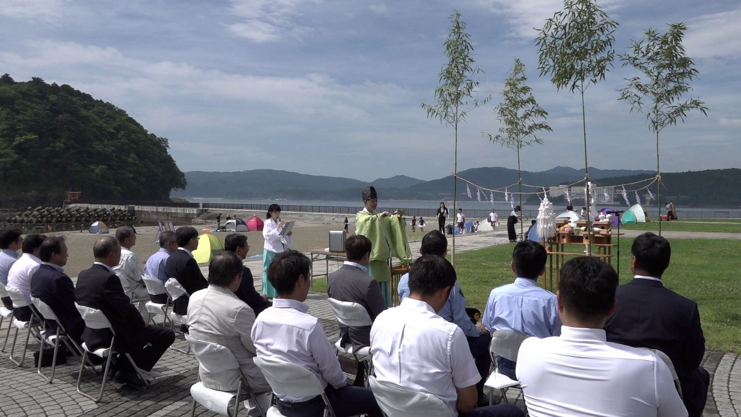 海辺の芝生広場で、多くのスーツ姿の男性たちが座って神職の祝詞を聞く中、海を背景に笹竹としめ縄、供え物が並ぶ神事が行われ、晴れた空の下で式が進んでいる写真