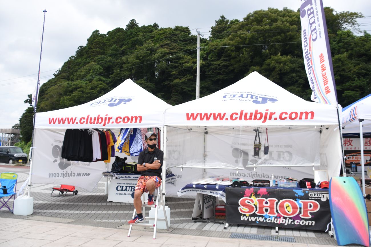 白いテントの下で、黒いマスクと黒いシャツに赤い柄の短パンを着た男性が椅子に座り、手を組んで店舗ブースの前にいる写真