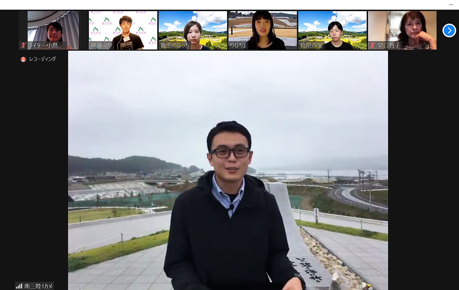 屋外の記念碑の前で男性がカメラに向かって話している様子を、複数の参加者がオンライン会議システムで見守っている写真