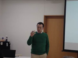 緑色のセーターを着た男性がペンを持った右手を挙げて話をしている写真