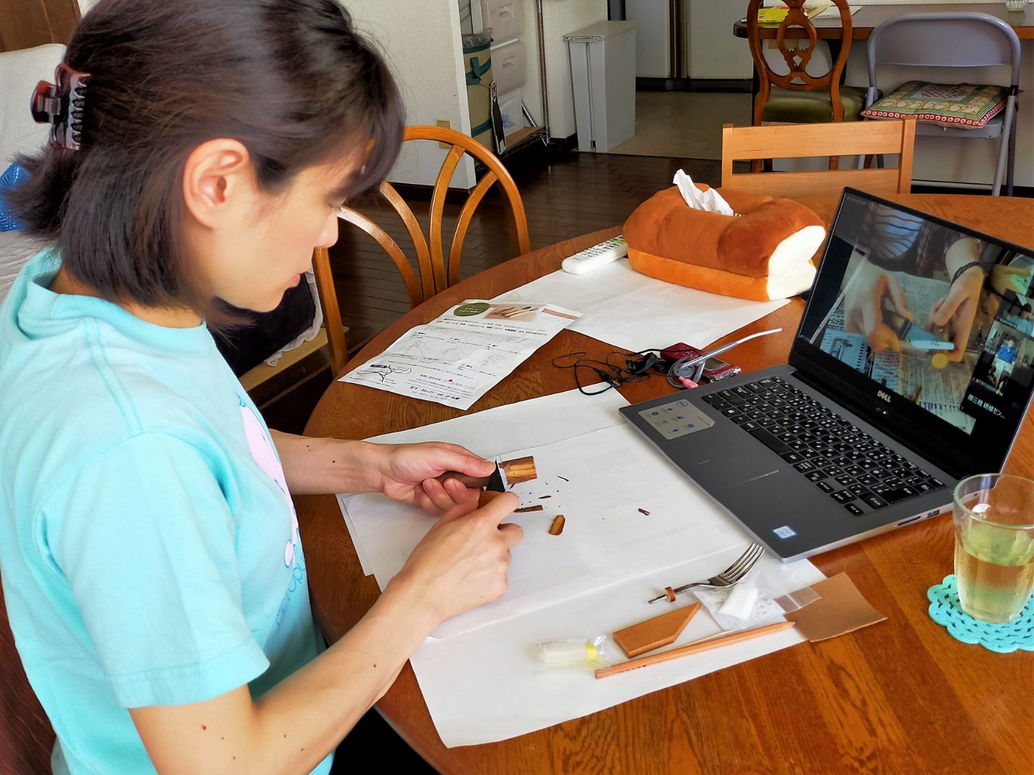 女性が自宅のテーブルでパソコンのオンライン映像を見ながら、ナイフで木の枝を削ってフォークの持ち手を作っている様子の写真