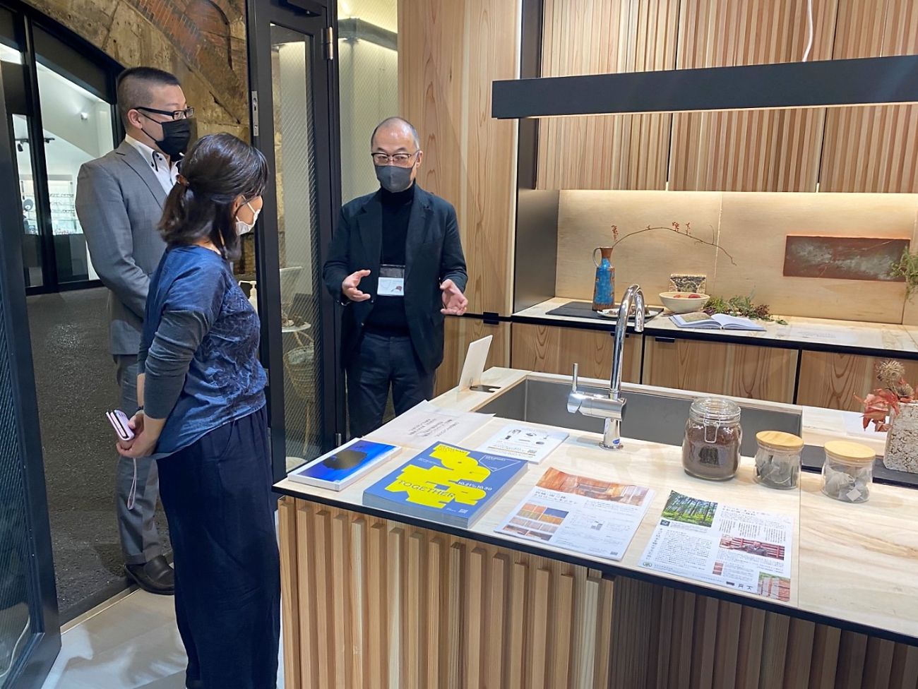 木目調のキッチンで、説明をするスーツ姿の男性と話を聞く二人の関係者が立っている写真