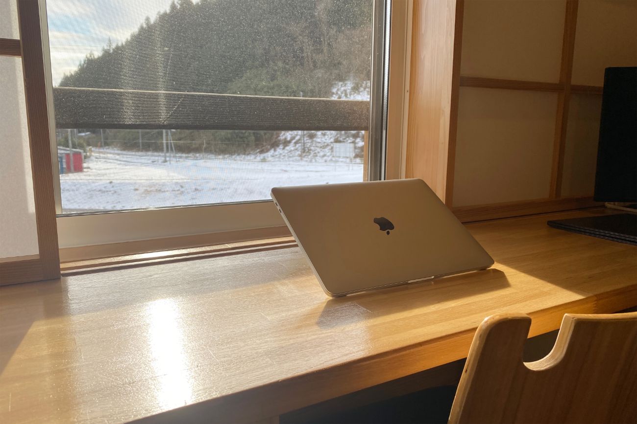 窓側の木の机の上に開いていないノートパソコンが置かれ、窓の外には雪景色と山が見える作業スペースの写真