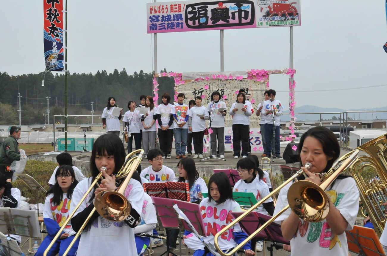 屋外ステージで高校生たちが「COLOR BAND」と書かれたTシャツを着て楽器演奏や合唱を行い、「復興市」と書かれた看板のもとでイベントを盛り上げている復興支援音楽イベントの写真