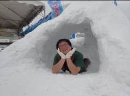 半袖のシャツの男性が、かまくらの中に寝そべって、両手で顔を支えて笑顔でポーズを取っている写真