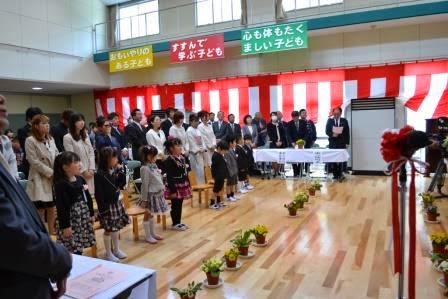 紅白幕が飾られた多目的ホールの窓側に先生方が立ち、中央に新入生の児童や保護者が立っている様子の写真