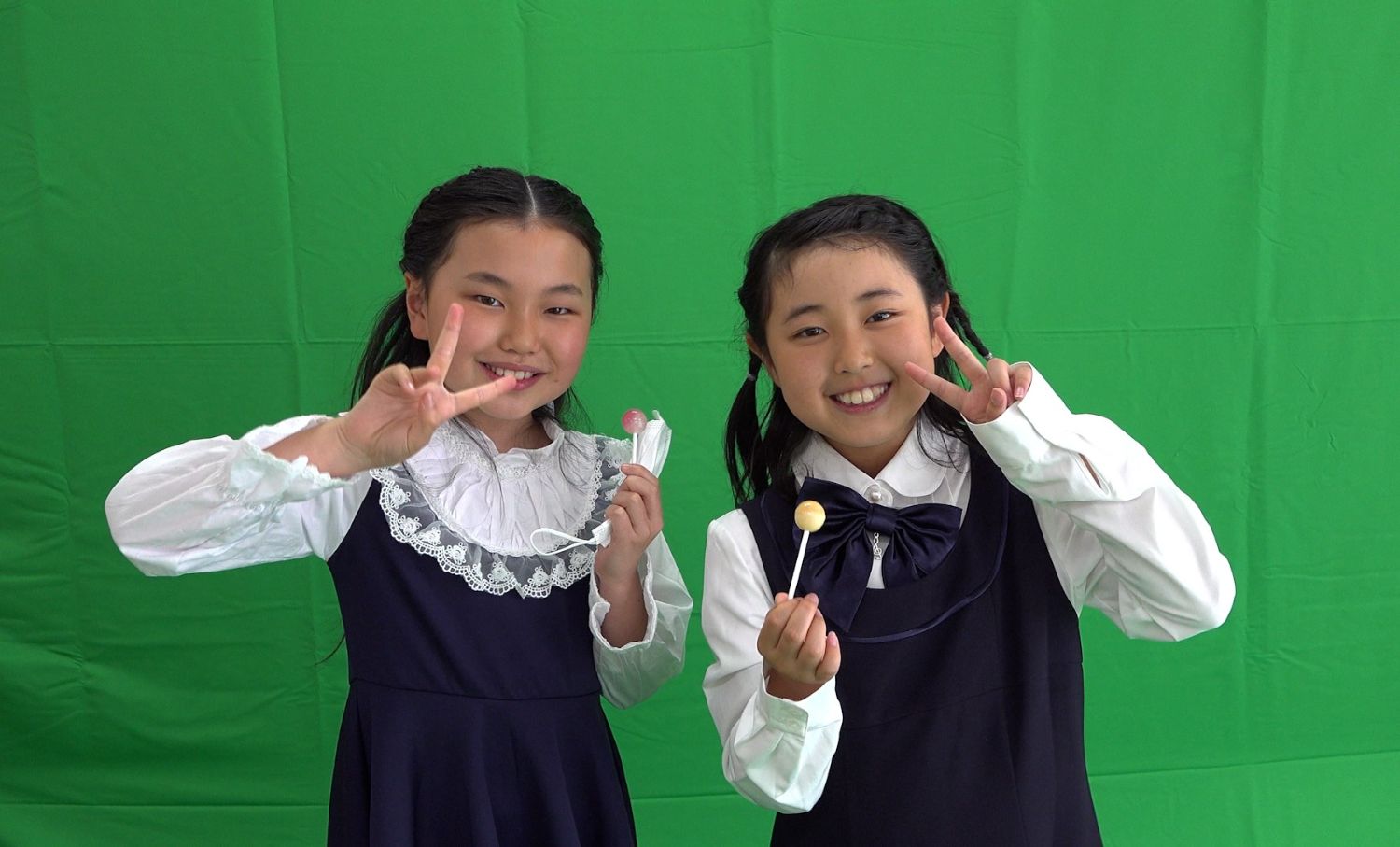 二人の女の子が緑色のクロマキー背景の前で、棒付きキャンディを持ちながら笑顔でピースサインをしている写真