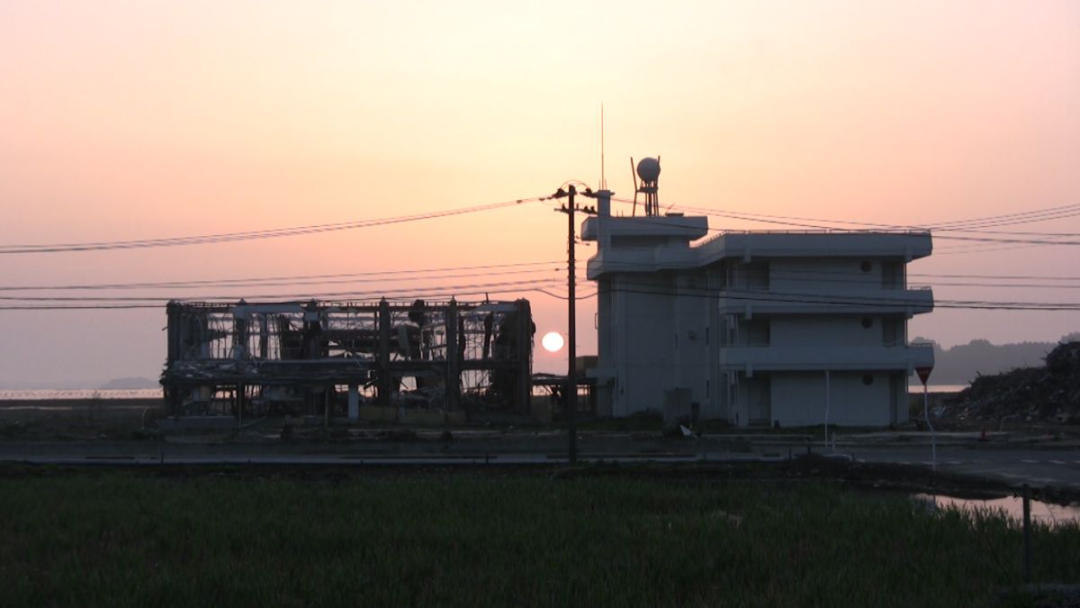 夕日が沈みかける中、左側に骨組みだけが残った建物と右側に無傷の白い建物が並んでいる被災地の風景の写真