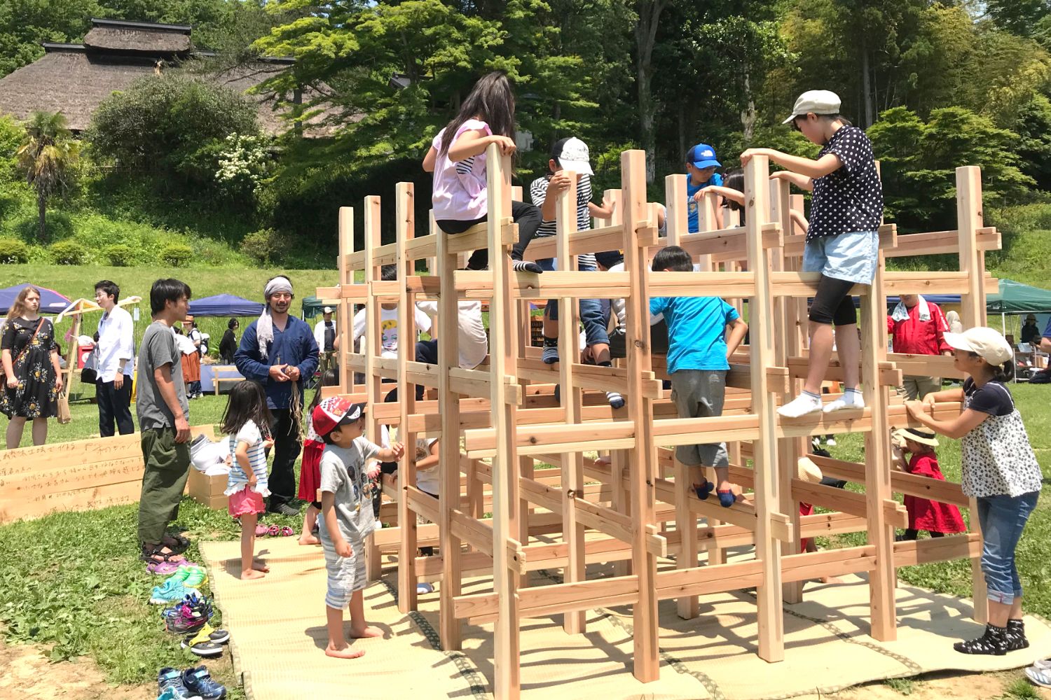 古民家や林背景に、広場に敷いたゴザの上に置かれた木材を組んで作ったジャングルジムのような遊具で子供たちが楽しそうに遊んでいる写真