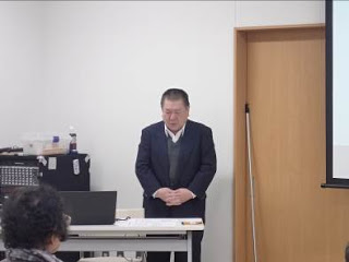 白い長机の後ろにスーツ姿の男性が立っている写真