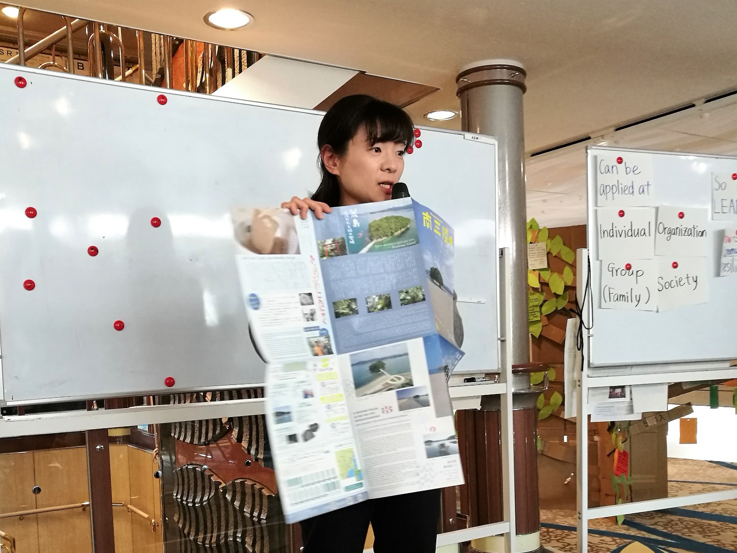 白板の前でマイクを持った女性が、南三陸町の情報が掲載された大きなパンフレットを広げて説明している様子の写真