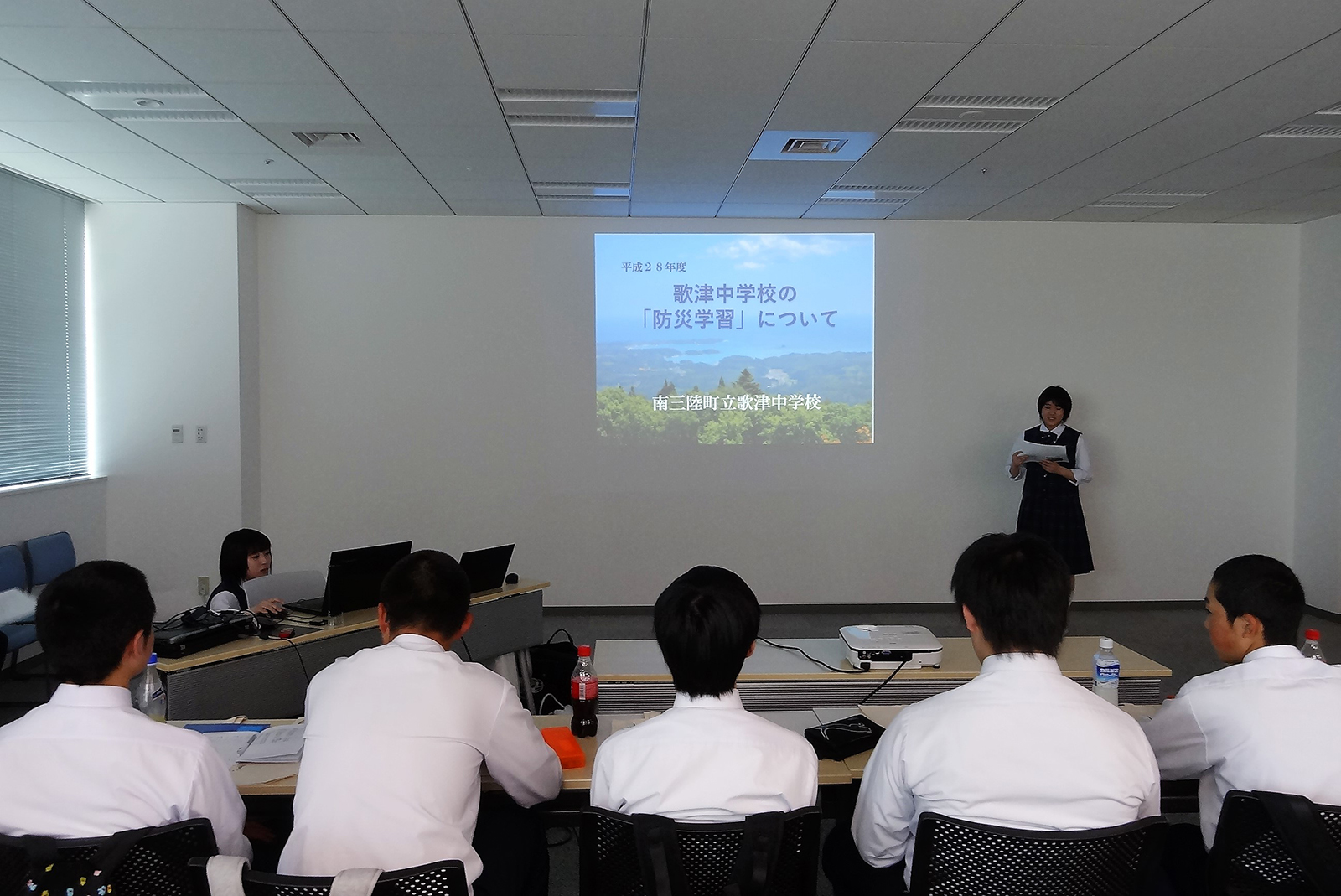 女子生徒が壁の前に立ち、壁に映し出された資料を使って発表を行い、他の生徒たちが着席して聞いている写真