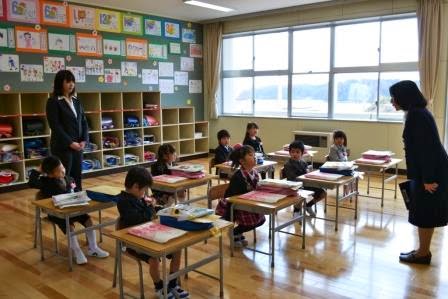 後方にスーツ姿の女性が立ち見ているなか、8人の新入生が2列に並んで座り、前に立っている女性教師の話を聞いている写真