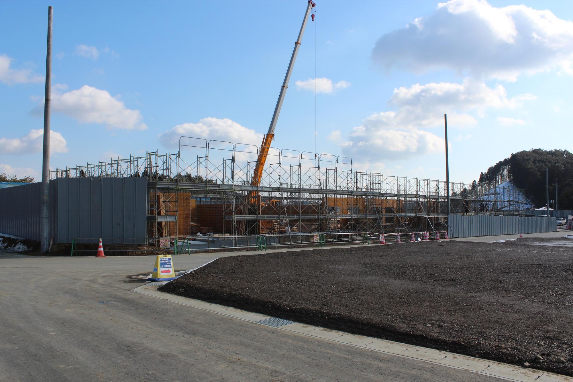 青空の下、鉄骨と足場が組まれた建設現場で黄色いクレーンが作業をしており、手前には舗装された道路と案内標識、奥には山林が広がる写真