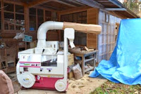 屋根のある木造の作業場の前に、白とピンクの籾摺り機（もみすりき）が設置されている写真