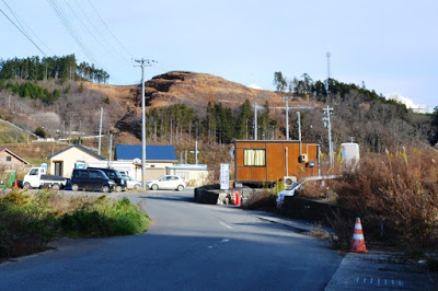 舗装された道路の先にプレハブ事務所や自動車が並び、枯れ草の生い茂る冬枯れの景色が広がる、奥に木々が伐採され山肌がむきだしになった小高い山で工事が行われている造成地の写真