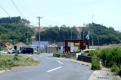 夏の青々とした緑に囲まれ、自動車やプレハブ事務所が並び、背景の山に工事の重機が見える、住宅地入り口の様子の写真
