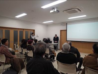 広い室内に並んだ椅子にたくさんの人々が座る中、天井に設置された大きなスクリーンの左側に男性が立っている写真