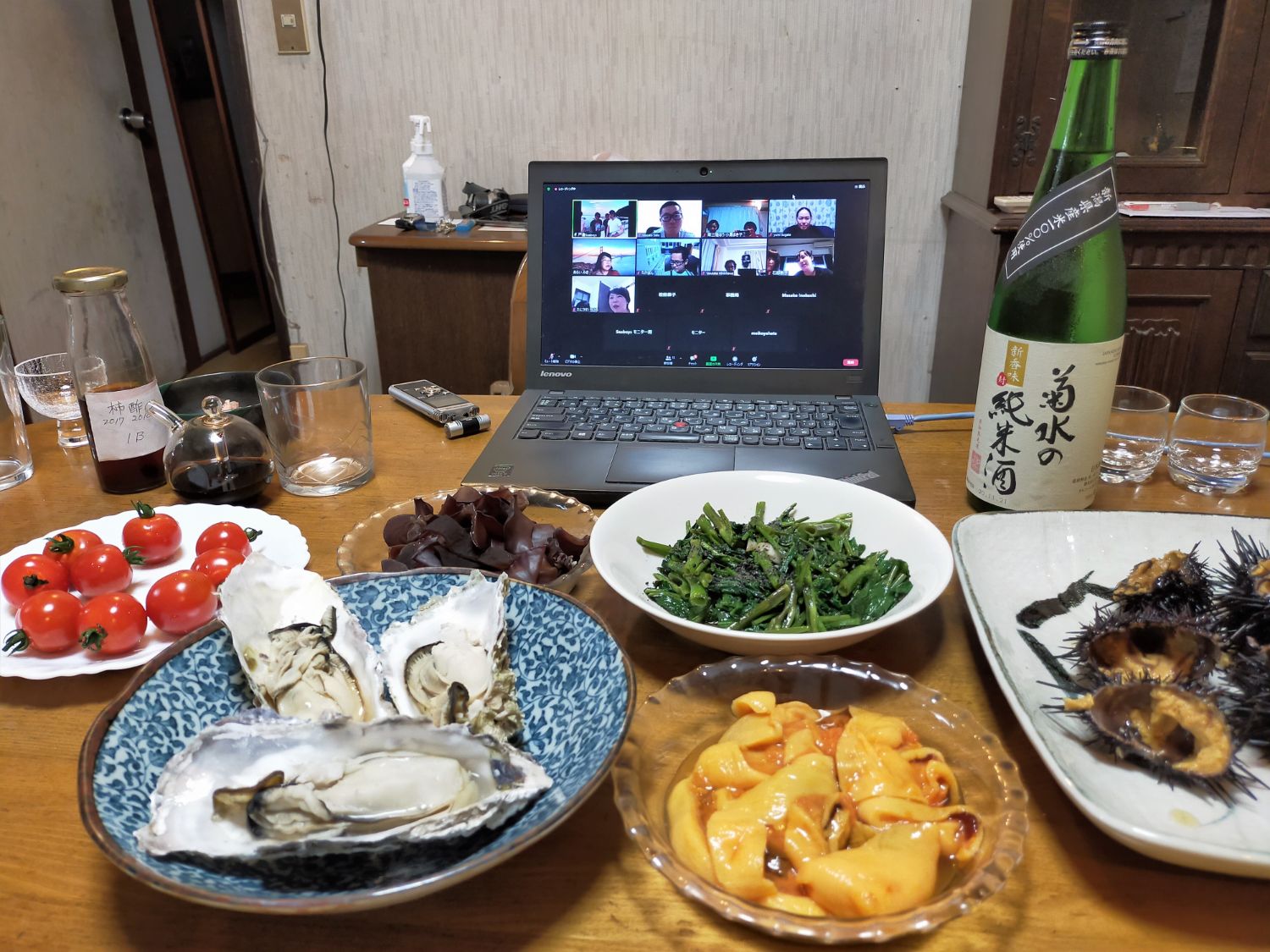 食卓の上に緑色の酒瓶、ミニトマト、ほうれん草のおひたし、蒸し牡蠣、ホヤ、殻付きウニなどが並べられ、中央のノートパソコン画面にはオンライン会議が表示されているオンライン食事会の写真