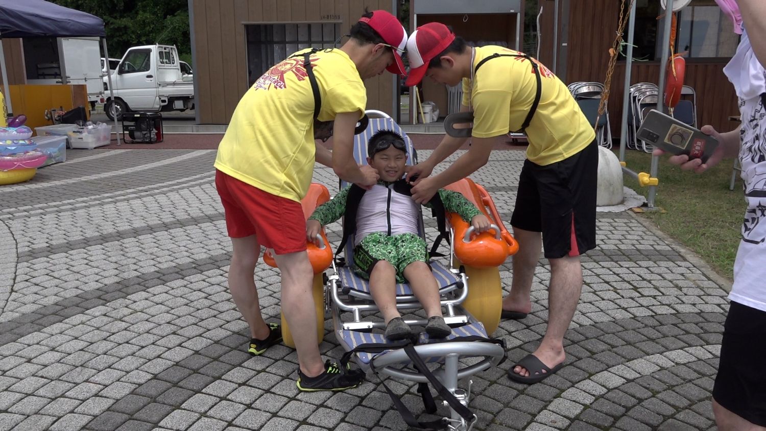 舗装された広場でライフガードのような黄色いシャツを着た男性2人が、水着姿の男の子をオレンジ色の浮き具付きビーチ用車いすに座らせてライフジャケットを装着している写真
