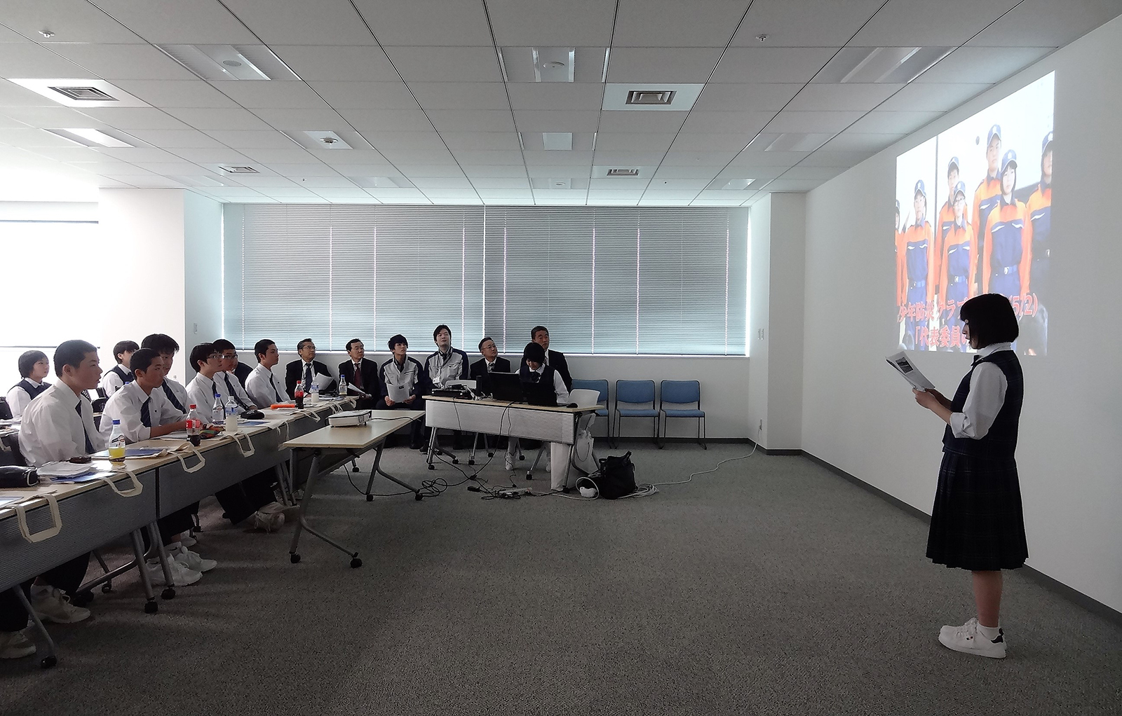 資料が投影された壁の前に立った女子生徒が、席に座っている他の生徒や関係者たちに向けて発表を行っている様子の写真