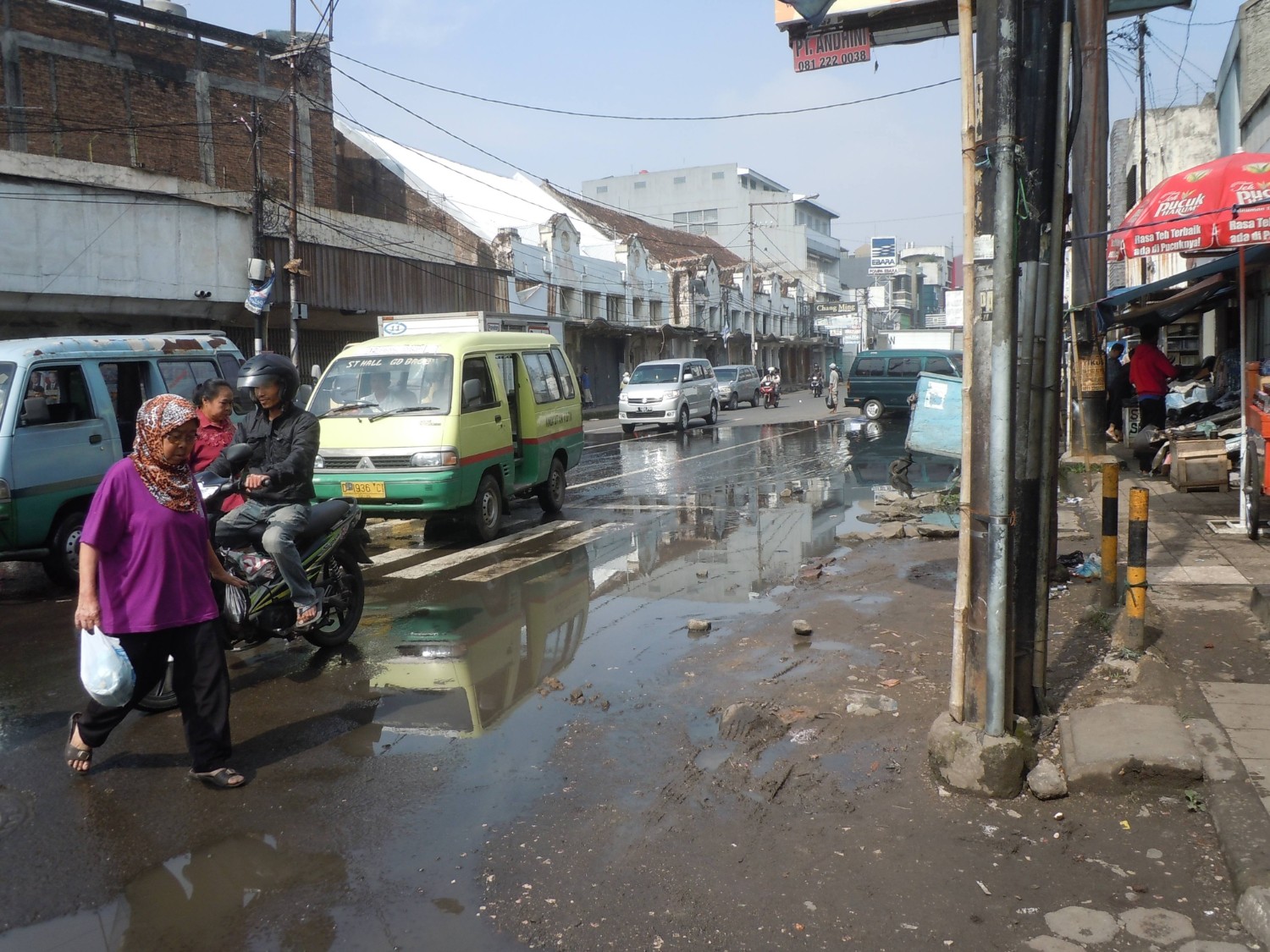 雨上がりで水たまりができた道路を、バイクに乗る人や歩いて渡る人々、ミニバスが行き交うバンコクの市街地の様子の写真