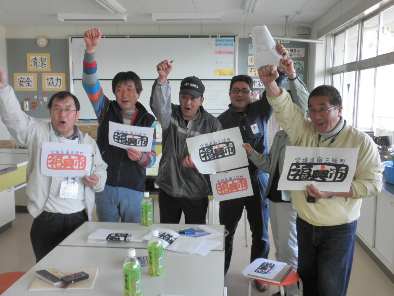 教室のような室内で、男性たちが「復興市」と書かれた紙を掲げて拳を上げ、笑顔で元気にポーズをとっている写真