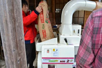 男性が、籾摺り機に袋に入った籾（もみ）を投入している様子の写真