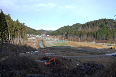 伐採された木が手前に積み重なり、中央を細い川が蛇行しながら流れ、周囲を森林に囲まれた広い造成地にオレンジ色の重機や作業車が点在する工事中の写真