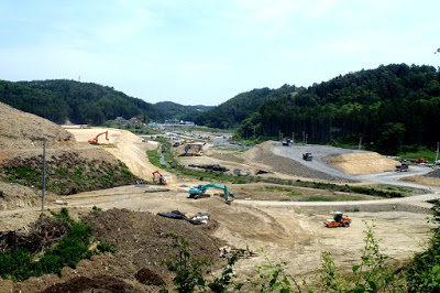 背景に緑の山々が見える広い谷間に造成された茶色い土の盛り土や舗装中の道路が伸び、複数のショベルカーやトラックが作業ている工事現場の写真