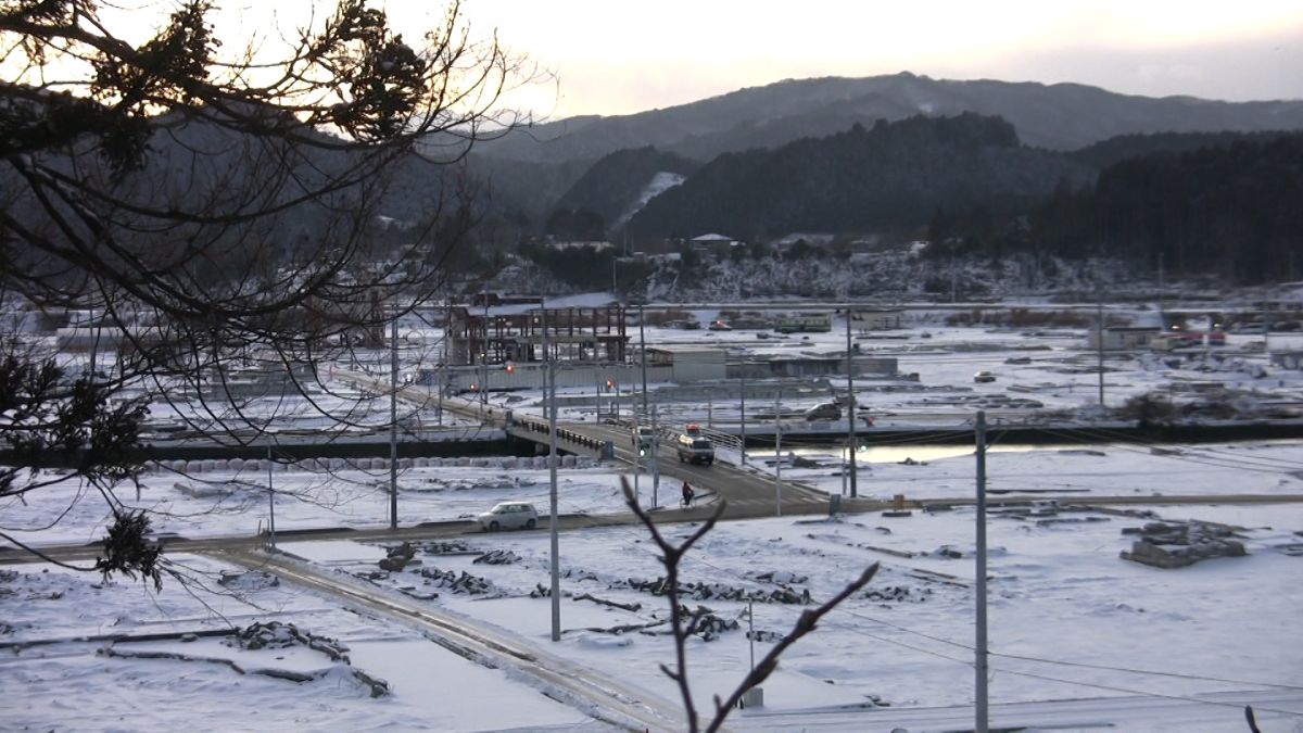 雪に覆われた山間の町を高台から撮影したもので、中央の川に橋が架かり、いくつかの車が行き交う復興の風景が広がっている写真