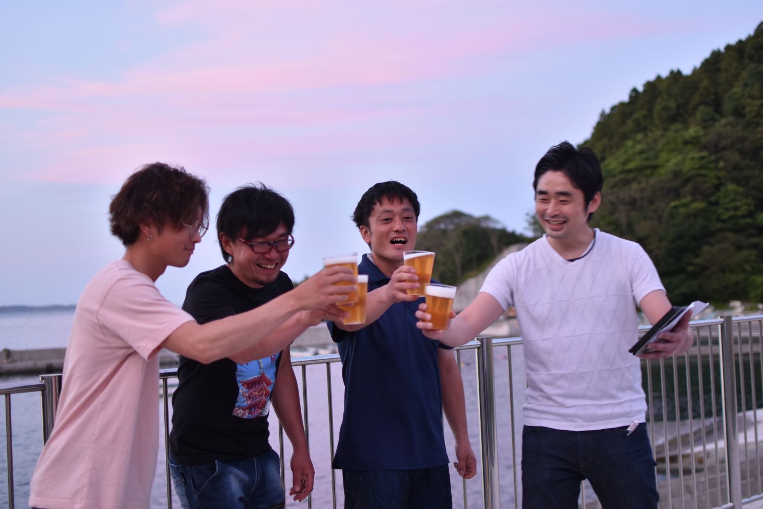 海辺の柵の前で4人の男性が夕焼け空の下でビールを手に持ち、笑顔で乾杯している写真