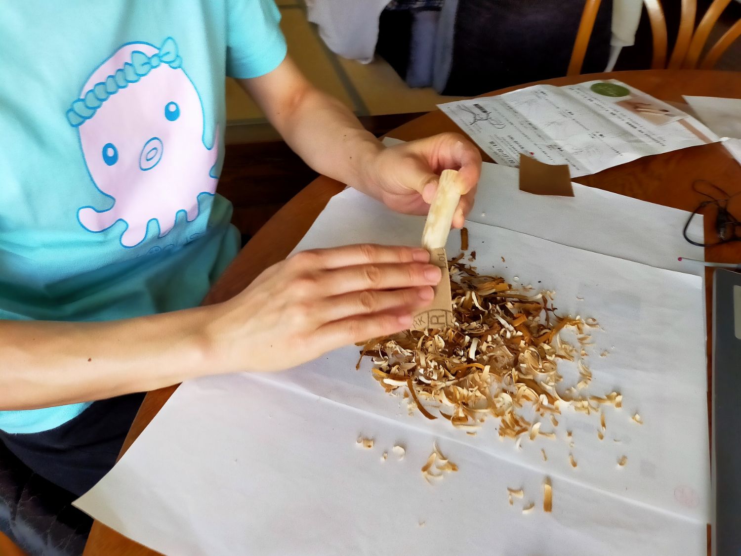 水色のタコのイラスト入りTシャツを着た人が、テーブルの上で木の枝を紙やすりで磨きながらカトラリーの持ち手を作っている様子の写真