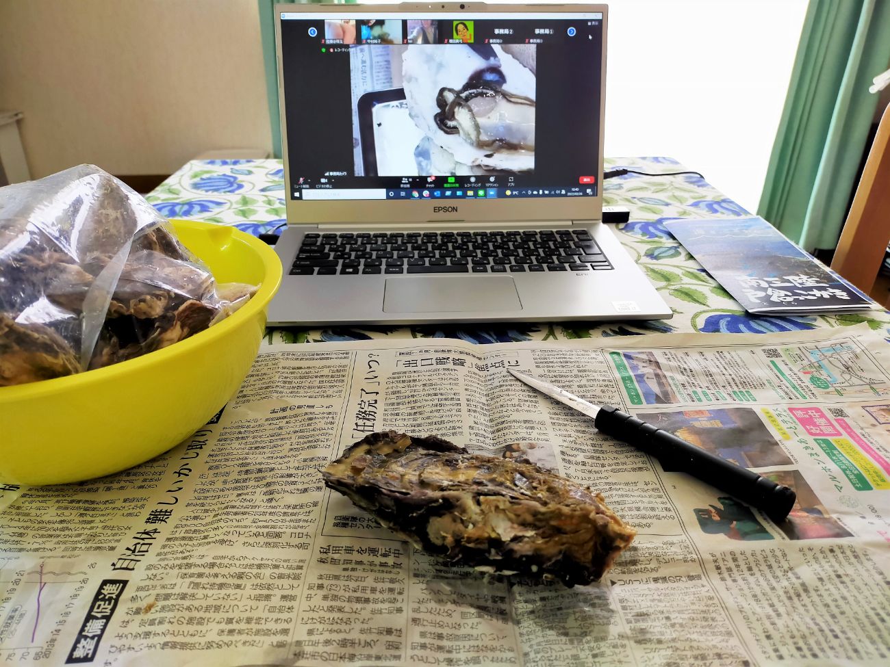 パソコンにZoomで牡蠣剥きの様子が映っており、新聞紙が敷かれた手前のテーブルには、牡蠣の殻、ナイフ、そして牡蠣が入った黄色いボウルがある写真