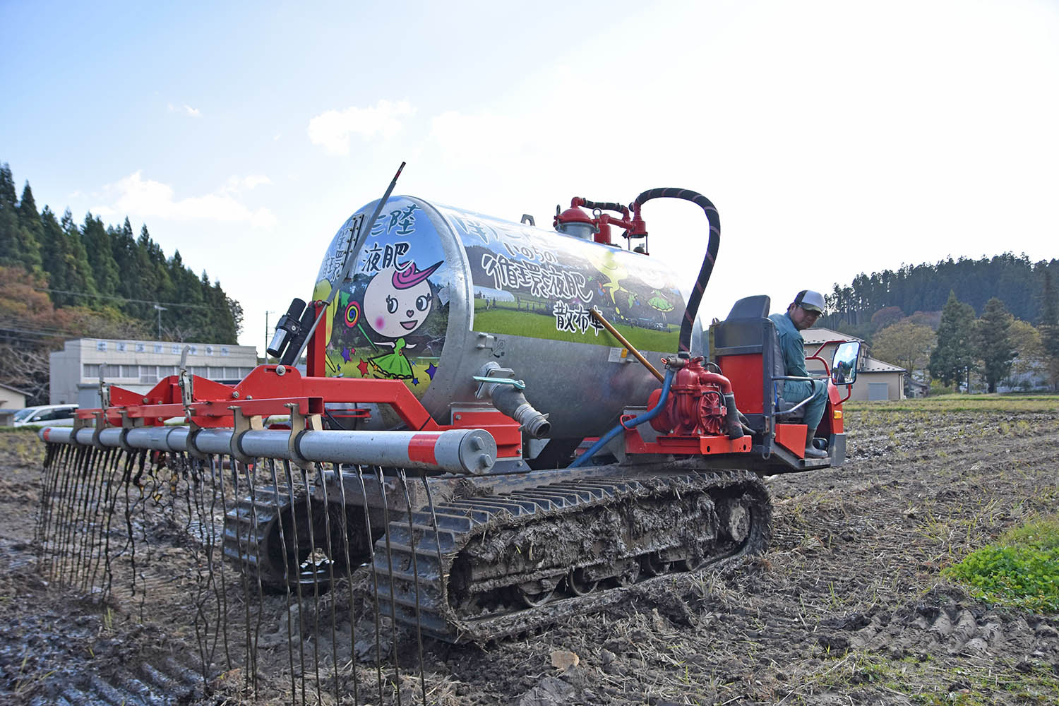 南三陸町の田んぼで、銀色のタンクにカラフルなキャラクターが描かれた農業用機械が、液体肥料を土に注入しながら進んでおり、運転席には作業着姿の男性が座って操作している様子の写真
