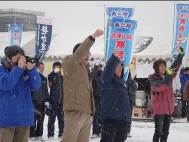 雪が降る中、テントや旗が立った会場に集まった来場者たちが、一斉に右手を挙げ、掛け声を上げている様子の写真