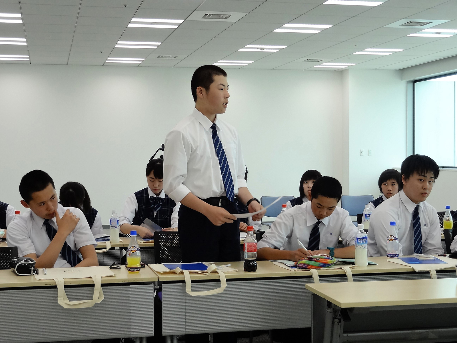 他の生徒たちが着席する中、1人の男子生徒が資料を手に立ち上がり、発言している様子の写真