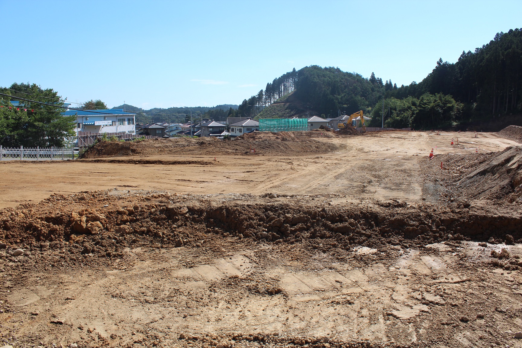 青空の下、広大な造成地の中で重機が作業を行っており、背景には住宅地や木々に覆われた小高い山が見える写真