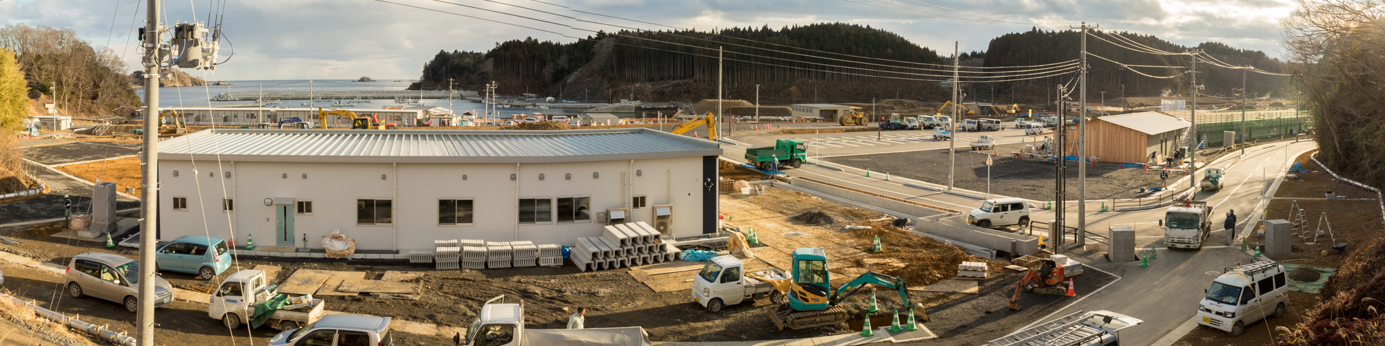 中央には白い建物があり、その周囲には複数の車両や重機が配置されている建設現場で背景には海と山が広がっているパノラマ写真