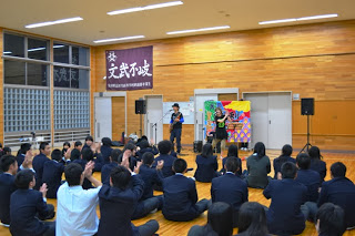体育館で座っている多くの学生が手拍子しながら前方で演奏する桃梨のライブを楽しんでいる様子の写真