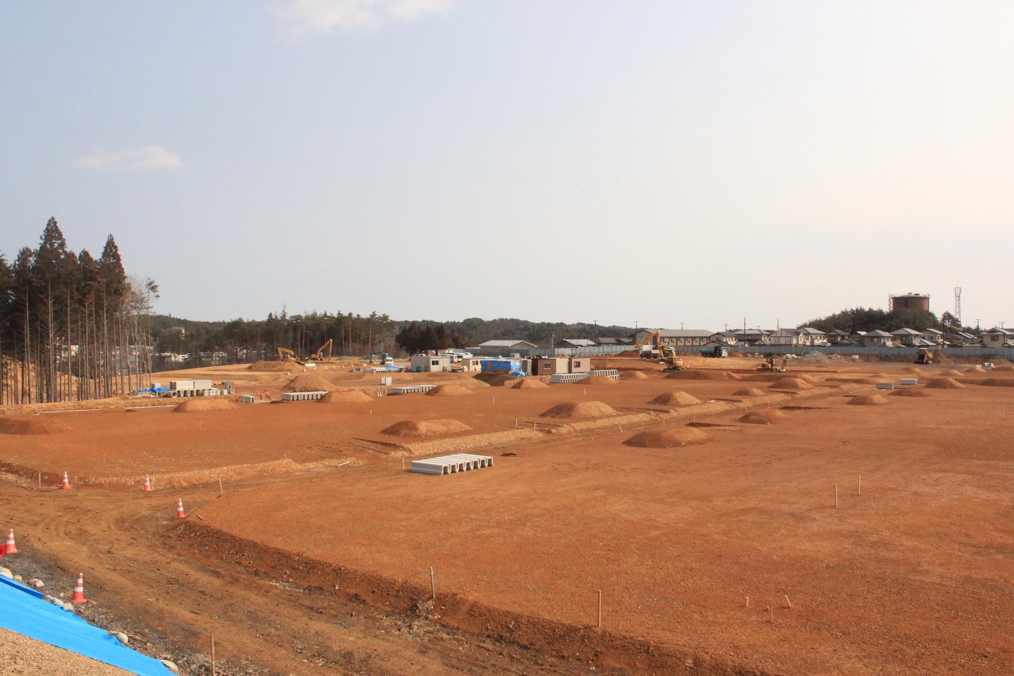 広大な赤土の敷地にいくつかの盛り土や建設機械が点在し、背景には木々や建物が見える様子を写している写真