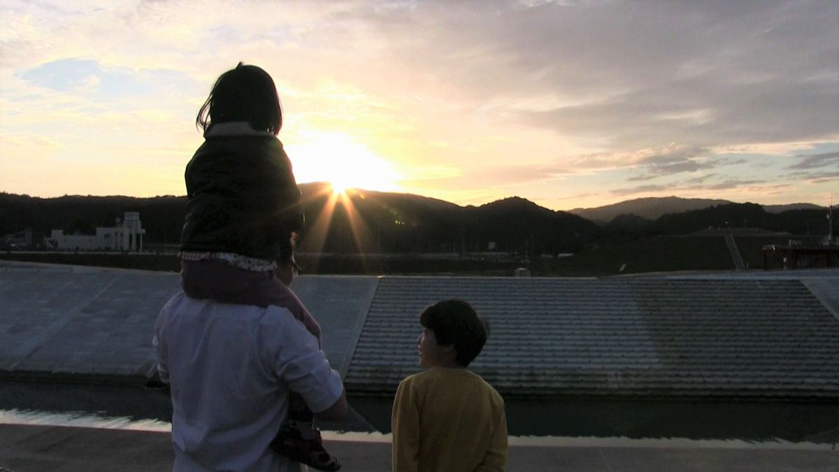 夕日が沈む川辺の堤防の上で、男性が肩車をした子どもともう一人の子どもとともに夕日を見つめる後ろ姿を捉えた写真