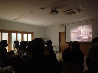 前方のスクリーンに映った映像を、席に座った参加者たちが見つめている写真