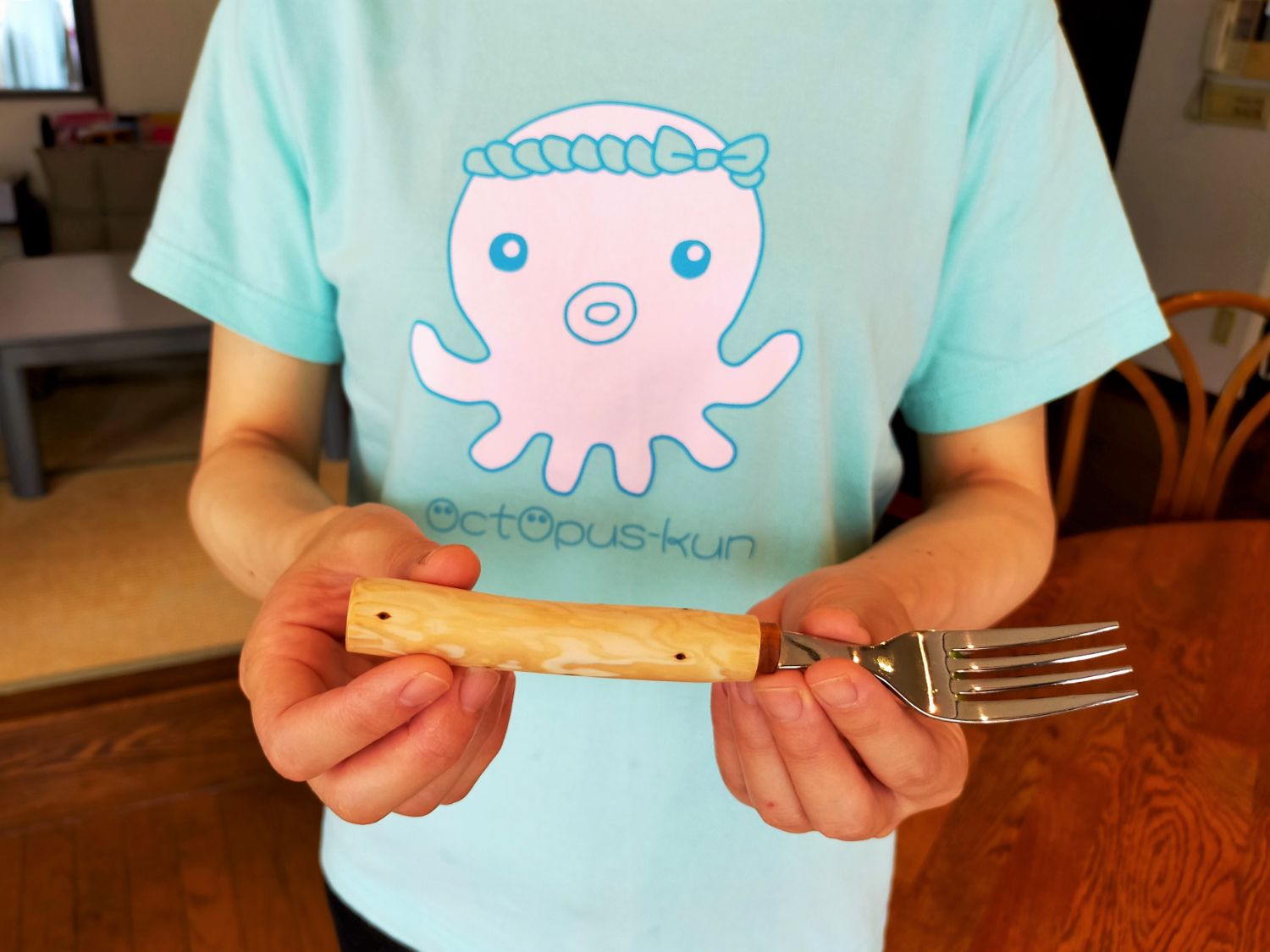 水色のタコのキャラクターが描かれたTシャツを着た人が、自作した木製ハンドルのフォークを両手で持っている写真