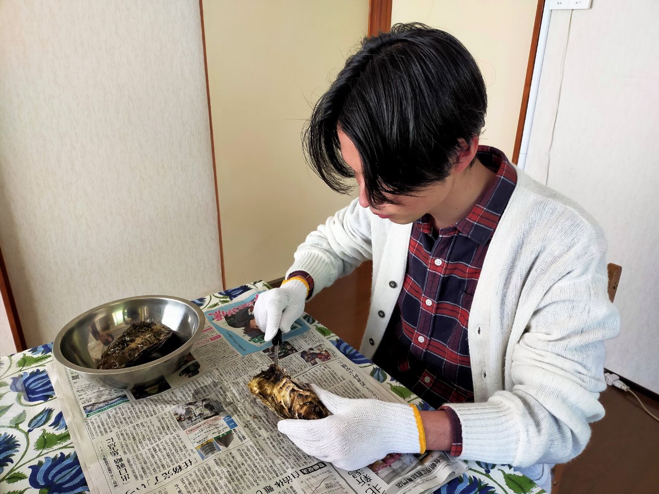 ステンレスのボウルや新聞紙が広げられているテーブルの上で、軍手をはめた手でナイフを使って牡蠣の殻をむいている男性の写真