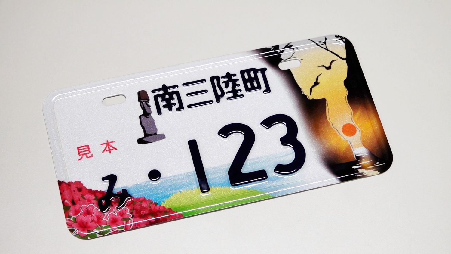 南三陸町のご当地ナンバープレートの完成品をクローズアップで写し、モアイ像や岩間から昇る朝日、つつじなどが描かれている、ナンバープレートデザインの完成品の写真