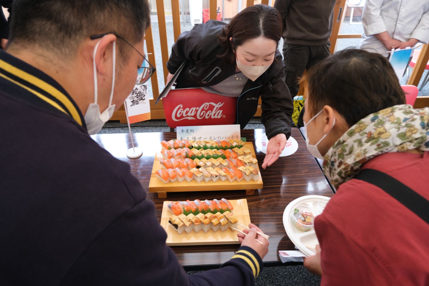 マスクをつけた3人がテーブルに並べられた握り寿司を見ながら選んでいる場面の写真