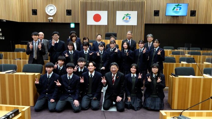 日本の国旗、市旗が掲げられた議場で、制服姿の中高生とスーツ姿の関係者の方々が整列してカメラに向かってピースサインをしながら笑顔で記念撮影している写真