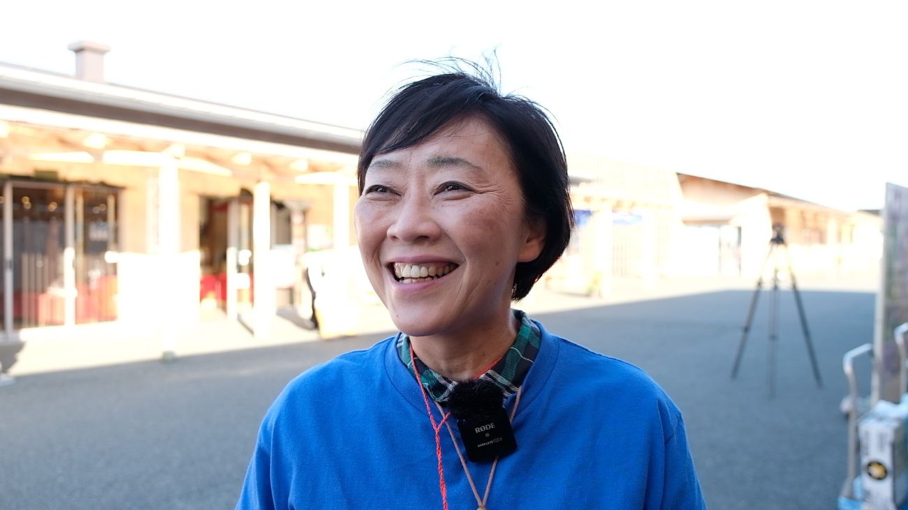 復興市の会場で青いスタッフTシャツを着た佐藤幸美さんが、笑顔でインタビューに答えている写真